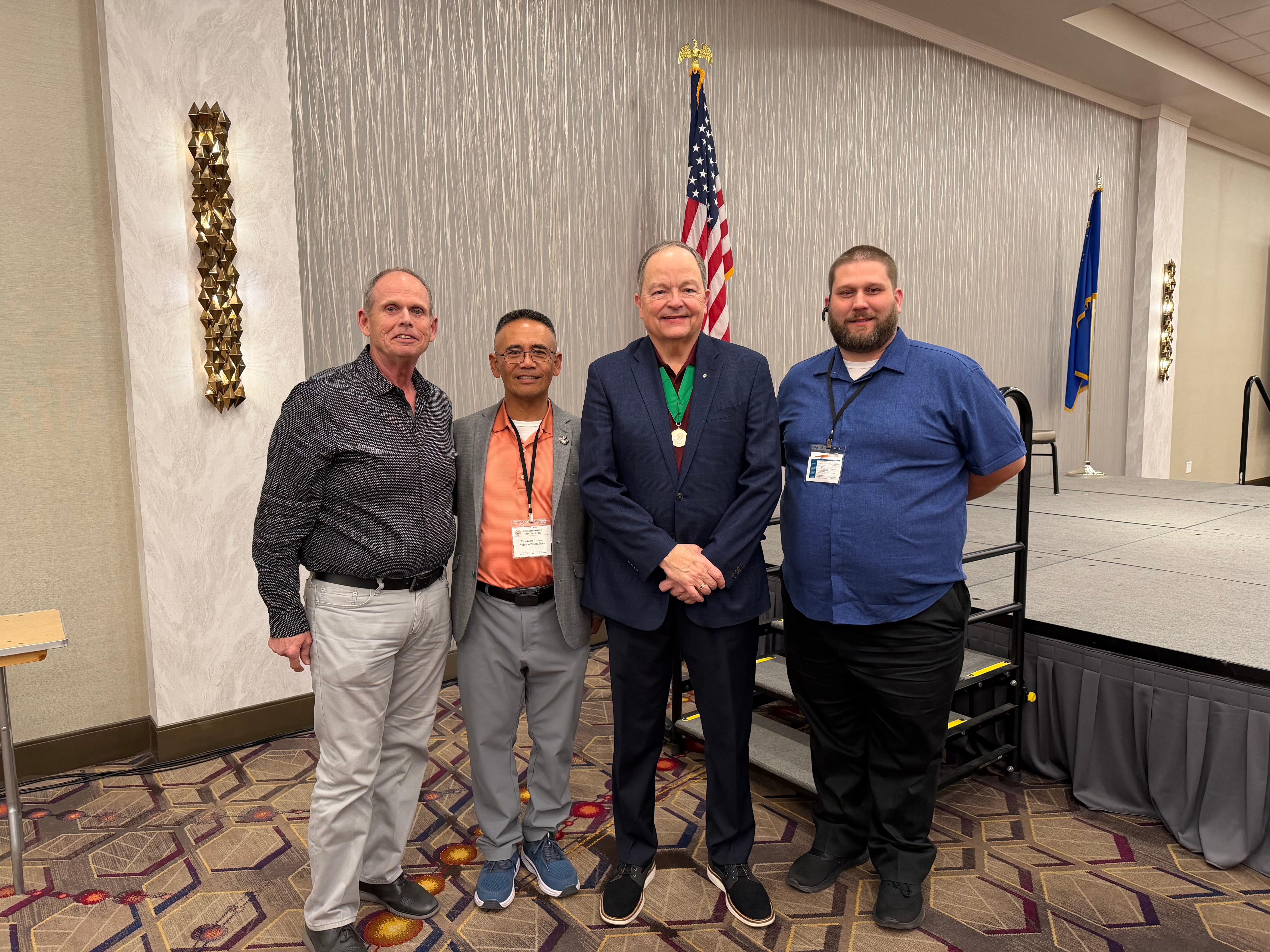 Four Scottish Rite members posing together at the Leadership Conference in Las Vegas, standing in front of American and state flags with a stage visible behind them